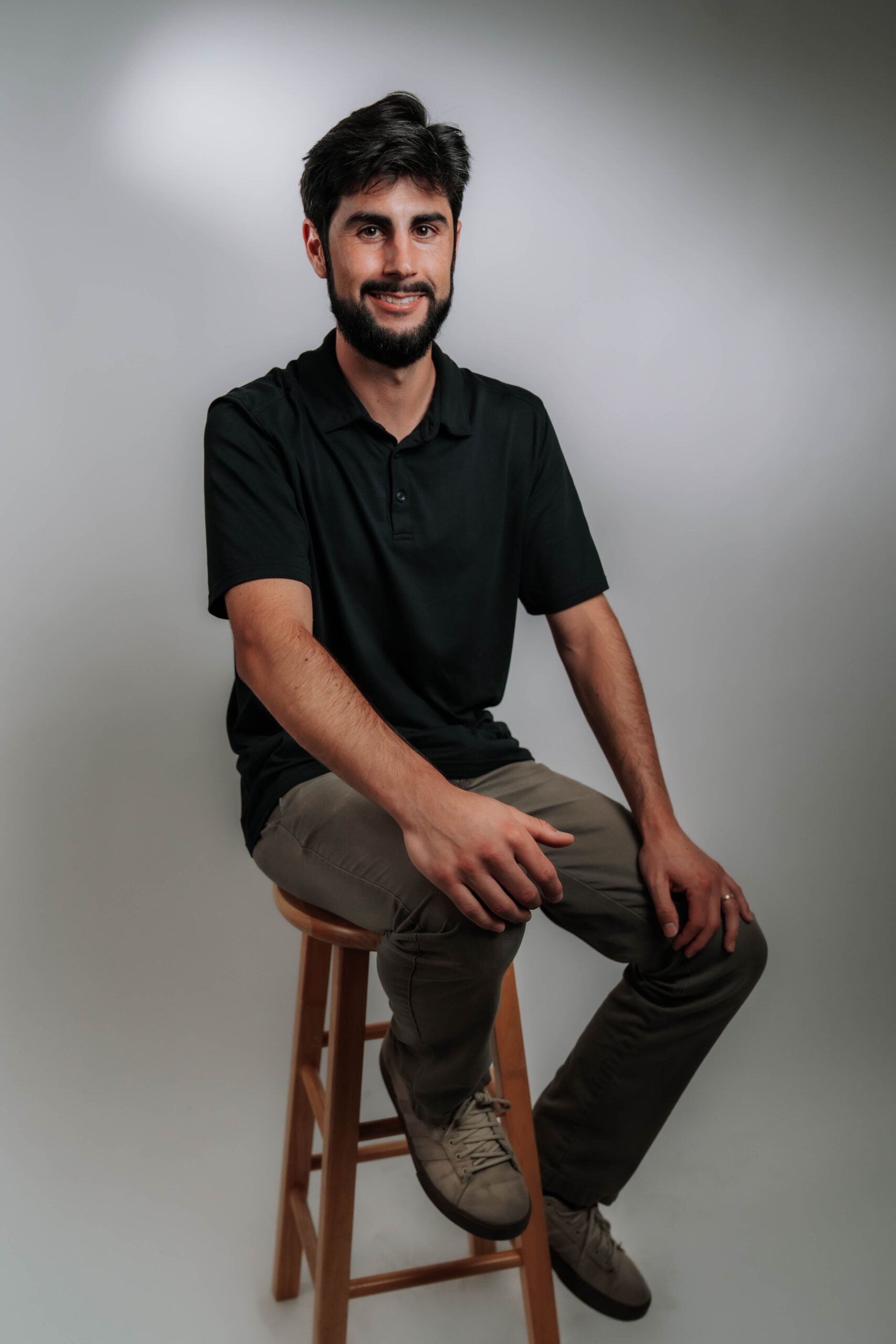 A personable professional headshot of Gaston Villanueva, Operations Lead at Advine Agency. Gaston's friendly smile and relaxed posture on a stool convey his approachable and effective leadership in optimizing agency operations.