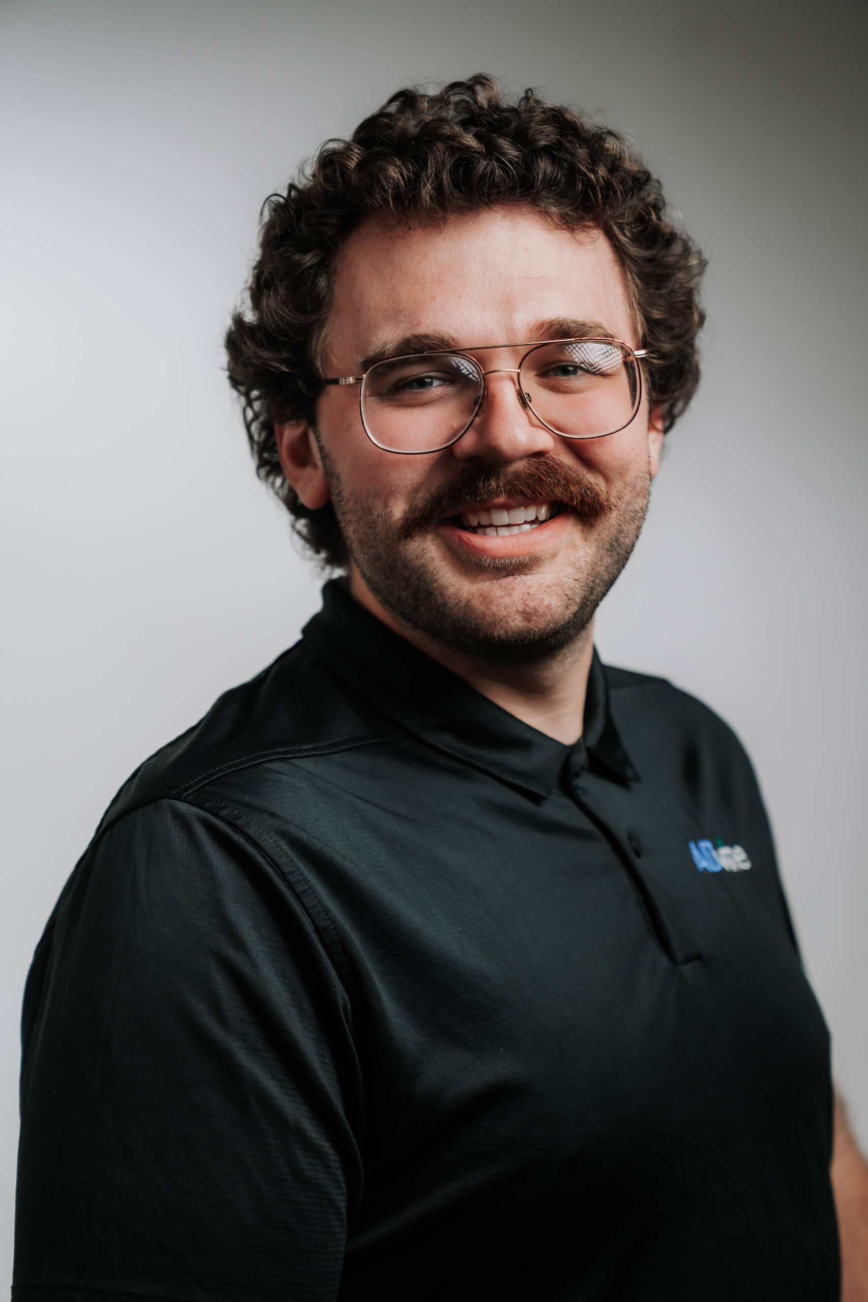 A charismatic professional headshot of John Pish, Multimedia Lead at Advine Agency. John's confident smile and professional attire, featuring a company-branded polo, reflect his creative vision and expertise in multimedia production.