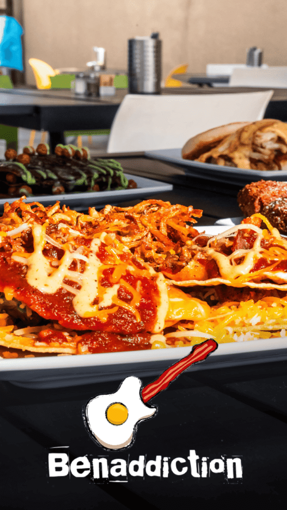 Benaddiction Advine's close-up shot of comfort food for Benaddiction captures a plate piled high with cheesy, saucy breakfast nachos topped with crispy fried onions, alongside other indulgent dishes, with the signature bacon-and-egg logo.