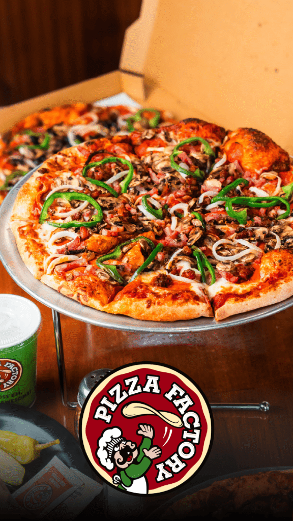 Pizza Factory Advine's food photography highlights a fresh, whole pepperoni and vegetable pizza on a metal stand for Pizza Factory, with a second pizza box visible in the background and a branded cup on the rustic wooden table.