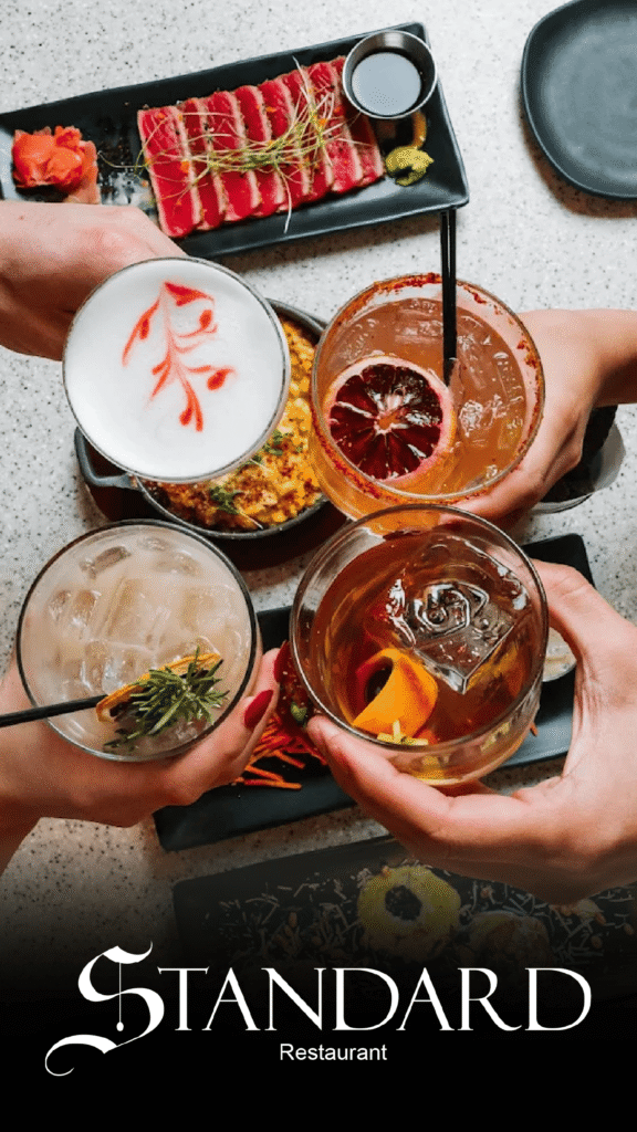 The Standard Advine's professional food and beverage photography captures a group of people holding four different, creatively garnished cocktails in a celebratory toast over a table set with sushi-grade tuna and other upscale dishes from The Standard Restaurant.