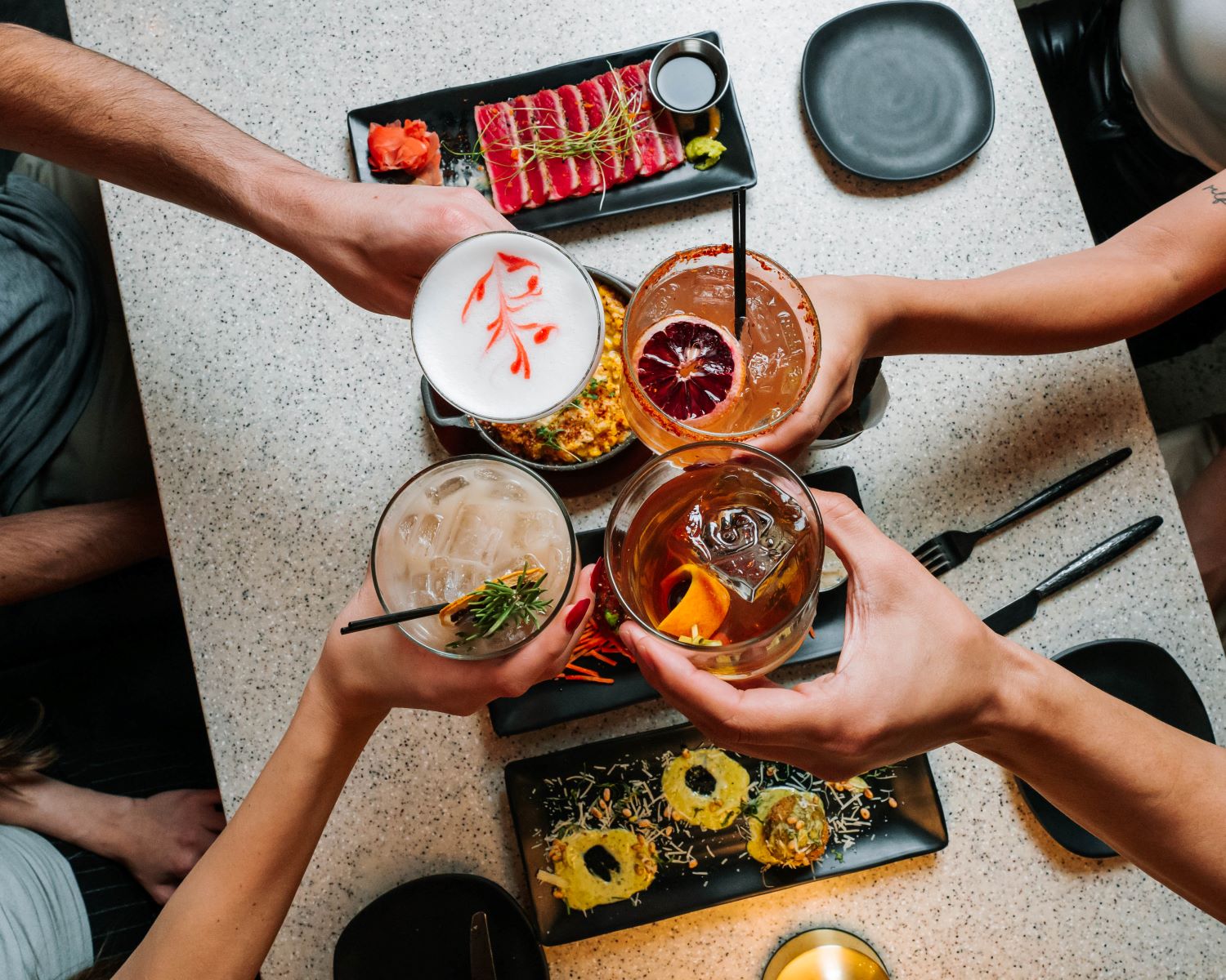 Four people are toasting with cocktails around a table laden with food. The table is light-colored and has various dishes, including what appears to be sliced tuna or beef, sushi rolls, and another dish with corn. Each person holds a different cocktail: one is a white drink with a red garnish, another is an orange drink with a blood orange slice, a third is a lighter-colored drink with ice and a sprig of rosemary, and the fourth is a dark amber drink with a twist of citrus peel. The hands of the people are visible, holding their glasses in the center of the frame, creating a sense of camaraderie and celebration.
