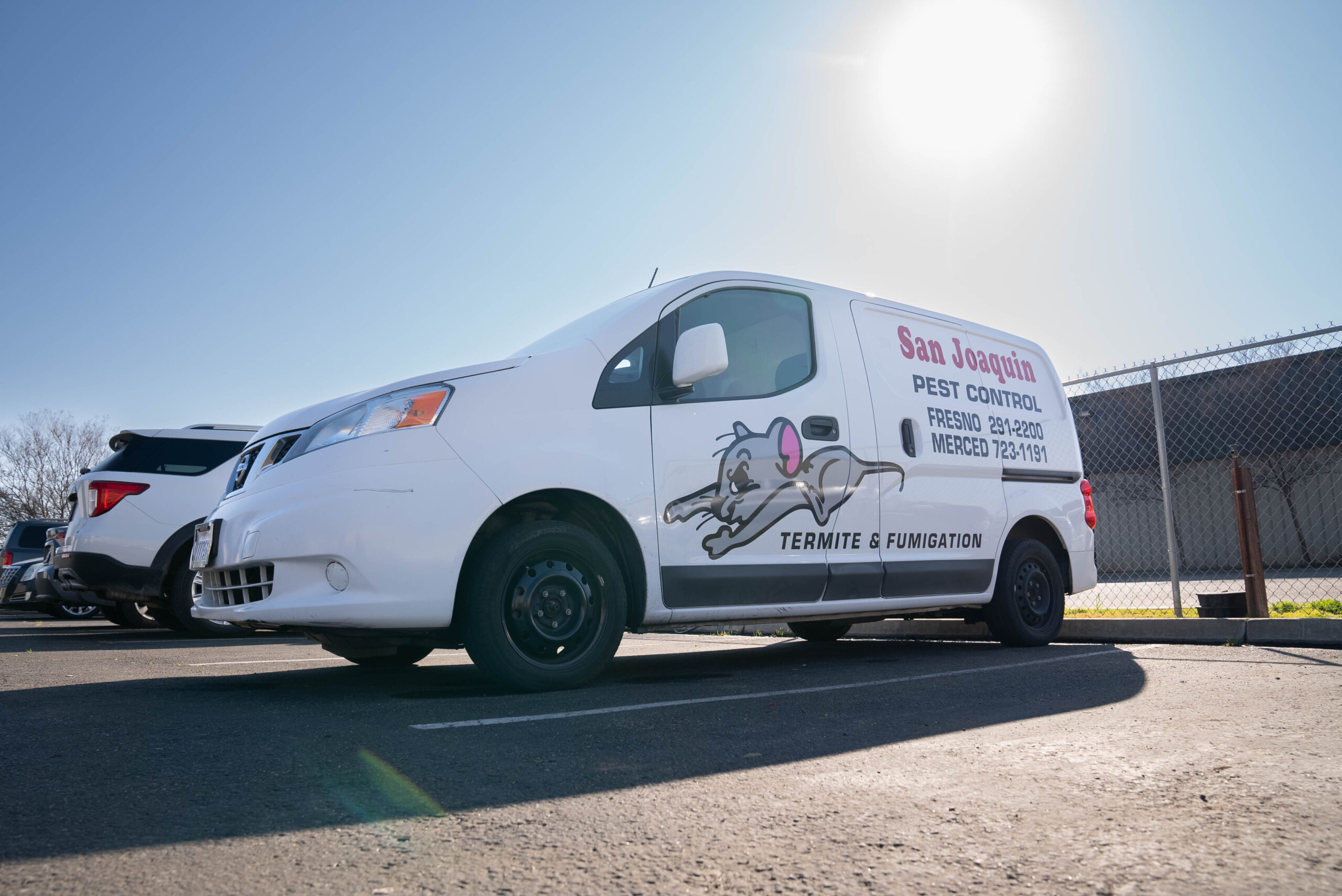 A white commercial van, possibly a Nissan NV200, is parked in a sunny outdoor lot. The side of the van features the company name and services in red and black lettering: 