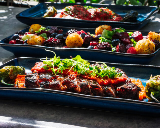 Group Food-1 "Three blue platters, expertly photographed by Advine's Photography Services in Fresno California, display a gourmet meal in natural light. The foreground shows a sliced, sauced steak with microgreens and blistered peppers. The middle platter features a vibrant salad of beets, berries, and golden croquettes,