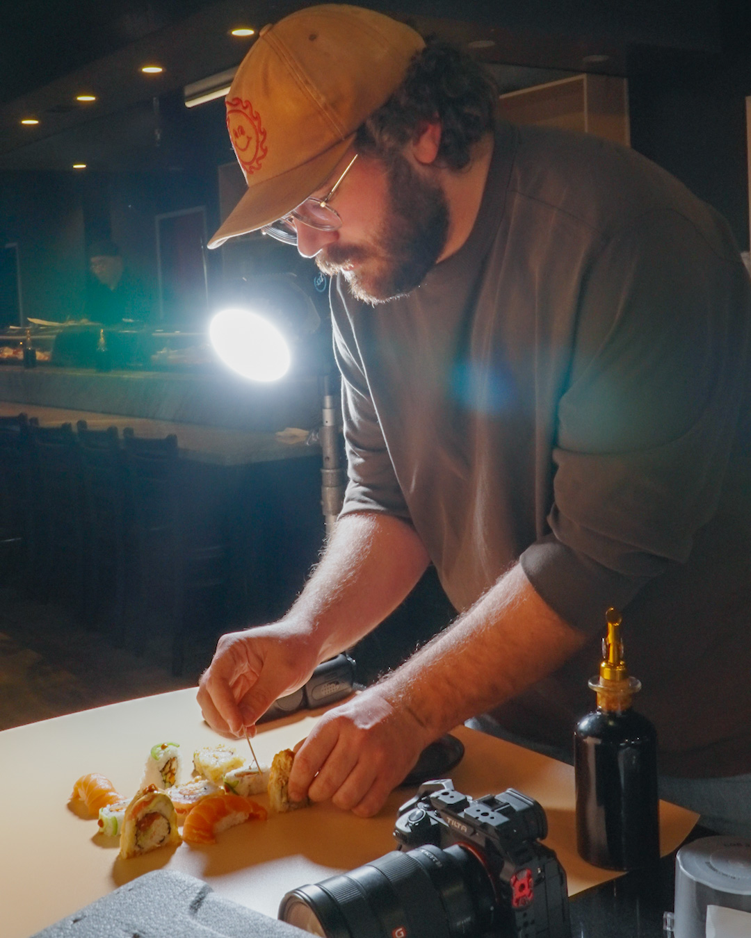 Behind-the-scenes shot of John, a professional food photographer from Advine's Photography Services in Fresno California, meticulously styling a row of sushi rolls on a light-colored surface. He is using a toothpick for precise adjustments, with a camera and lighting equipment visible in the foreground and background, demonstrating the expert process for high-quality commercial food photography.