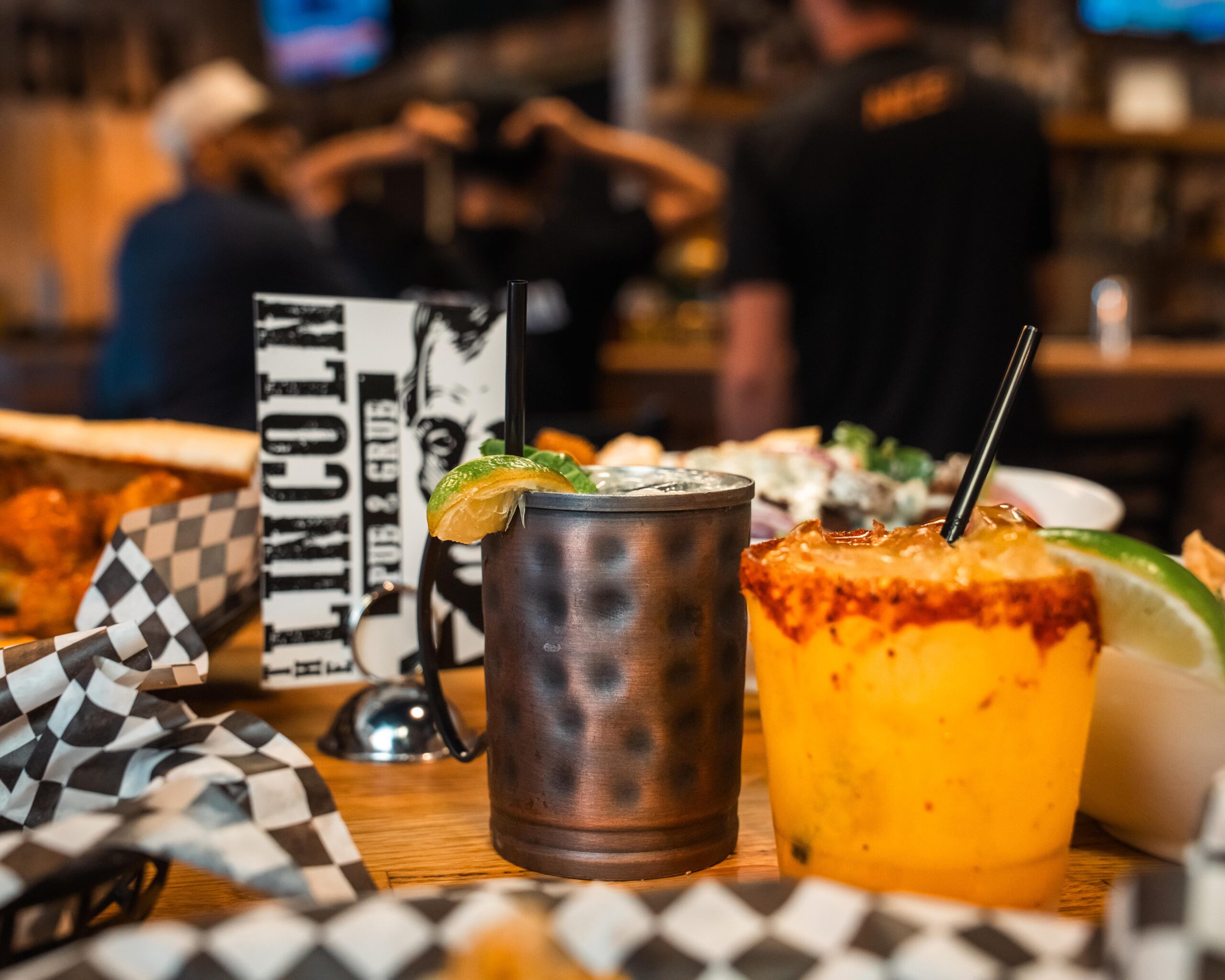 A close-up, low-angle shot of two cocktails on a wooden bar top or table in a dimly lit restaurant or bar, 