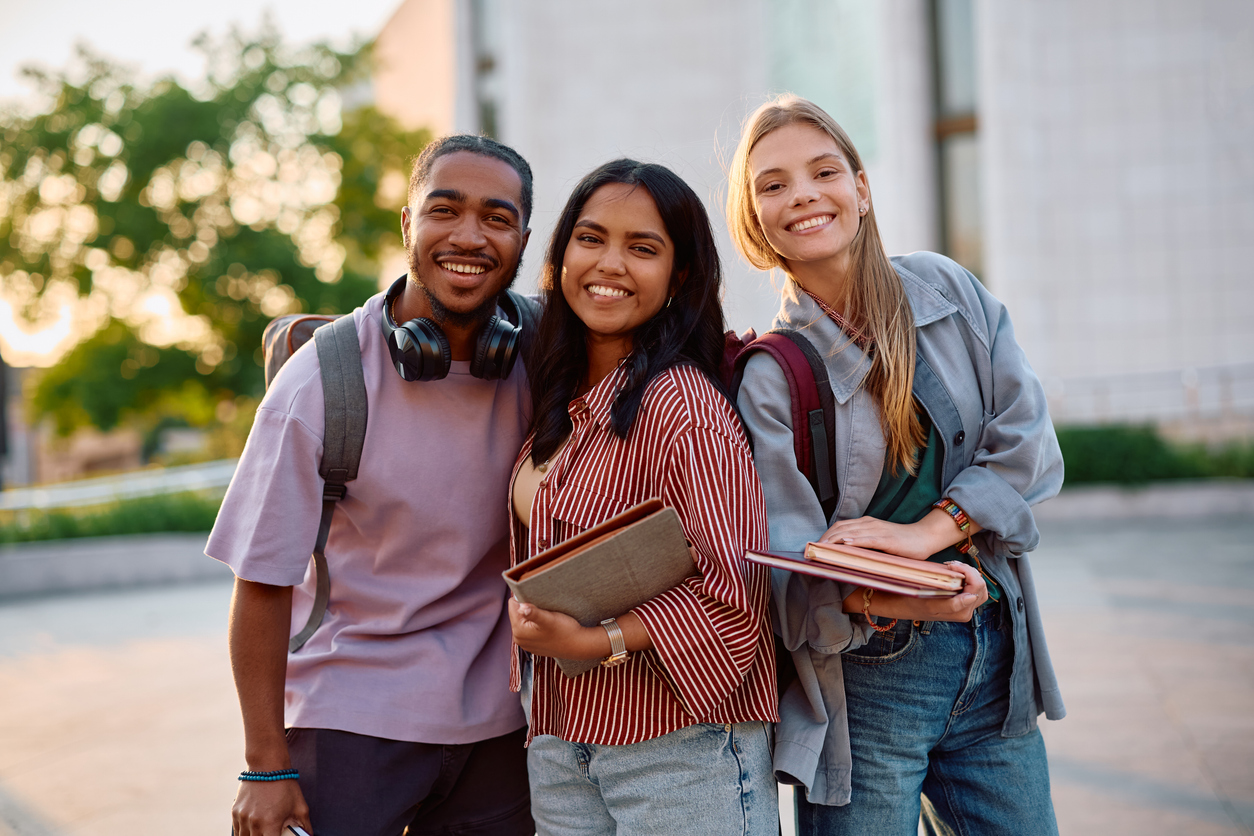 Group of happy students at university campus looking at camera. Three smiling college students, two women and one man, stand close together outdoors with backpacks on. They are casually dressed and holding books or notebooks. The man on the left is Black, wearing a purple t-shirt and headphones around his neck. The woman in the middle is South Asian, wearing a red and white striped shirt. The woman on the right is white, with blonde hair, wearing a gray jacket over a teal top and jeans. They all look happy and are facing the viewer. The background shows blurred greenery and a light-colored building, suggesting a college campus setting during the day.