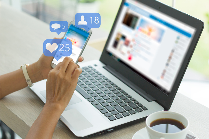 Social media engagement A person's hands are shown holding a smartphone displaying social media notifications for likes, comments, and new followers, indicated by overlaying icons and numbers (a heart with '25', a speech bubble with '5', and a person icon with '18'). In the background, a laptop with a blurred social media feed on the screen and a cup of coffee are visible on a wooden table. This image illustrates social media engagement, particularly relevant for local businesses tracking their online interaction and reach.