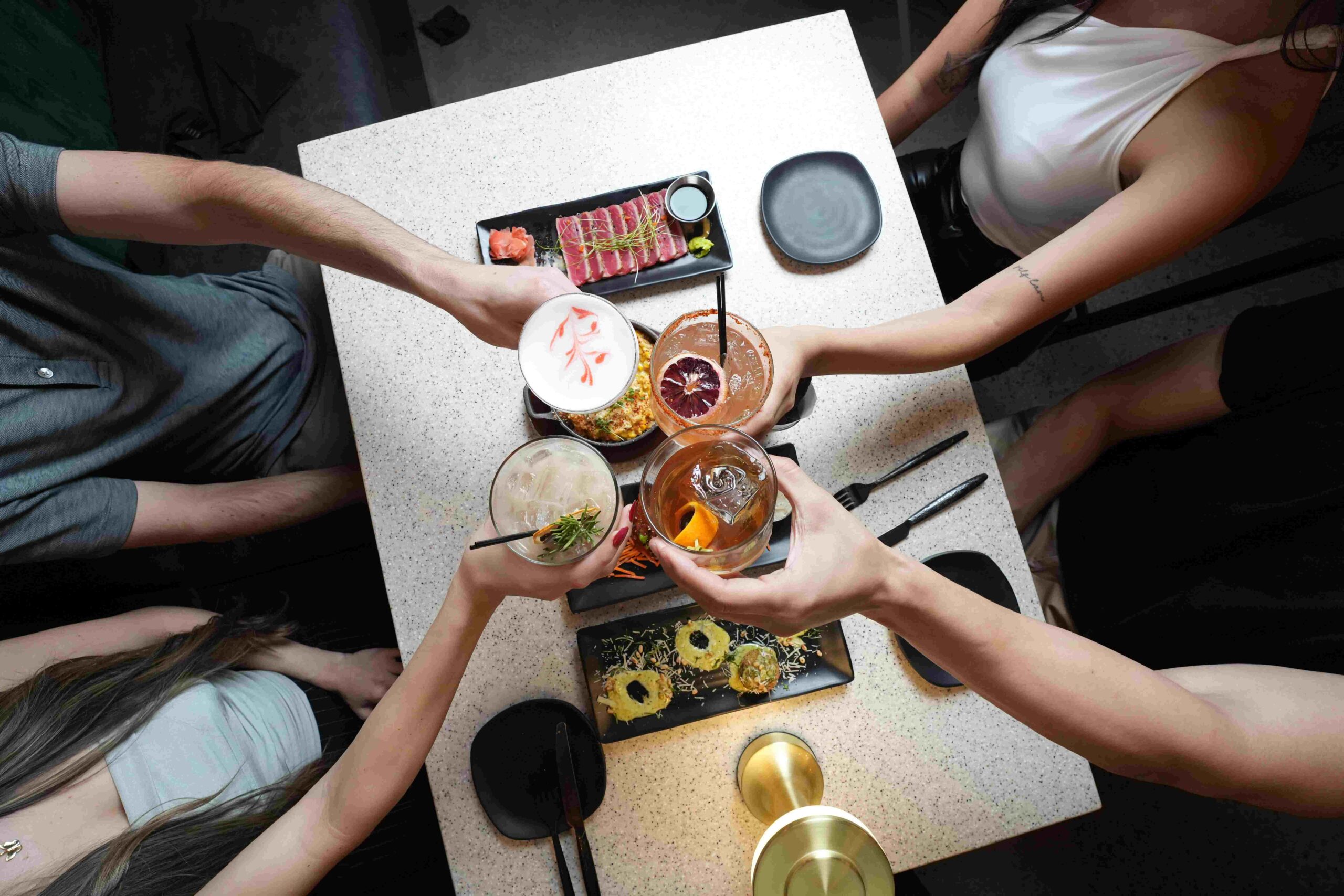 4 social media influencers cheersing their cocktails at a nightclub table full of appetizers and a warm table lamp