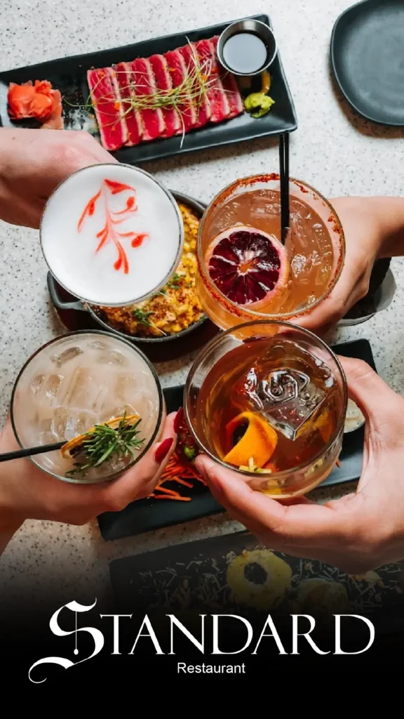 Advine's professional food and beverage photography captures a group of people holding four different, creatively garnished cocktails in a celebratory toast over a table set with sushi-grade tuna and other upscale dishes from The Standard Restaurant.