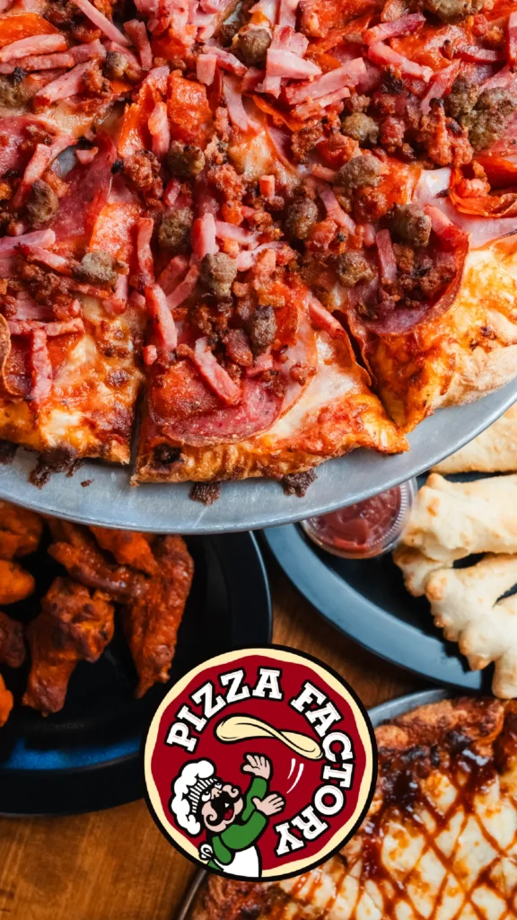 Advine's food photography highlights a fresh, whole pepperoni and vegetable pizza on a metal stand for Pizza Factory, with a second pizza box visible in the background and a branded cup on the rustic wooden table.